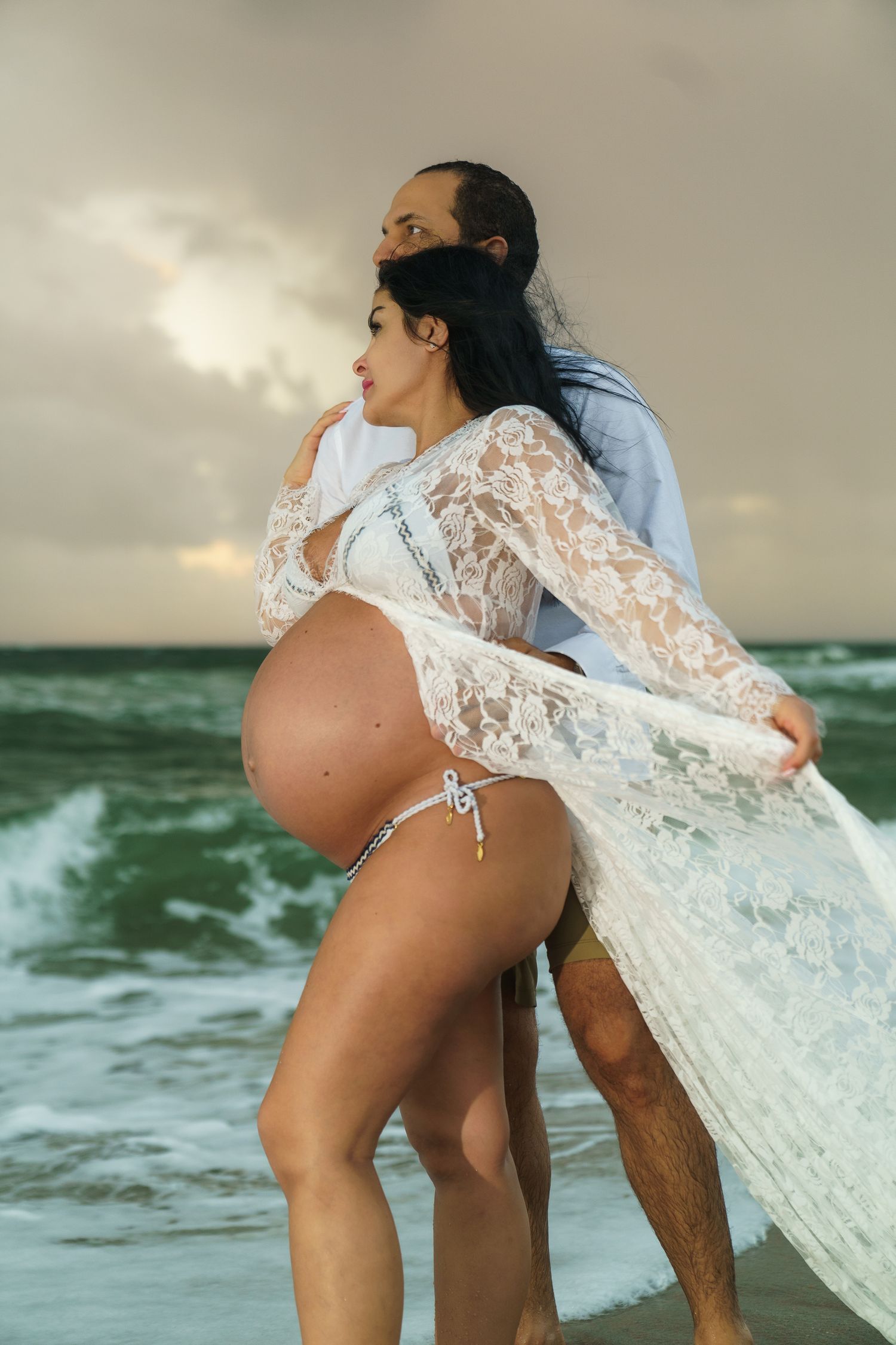 Couple holding baby bump during sunrise beach maternity session, golden hour, stormy skies