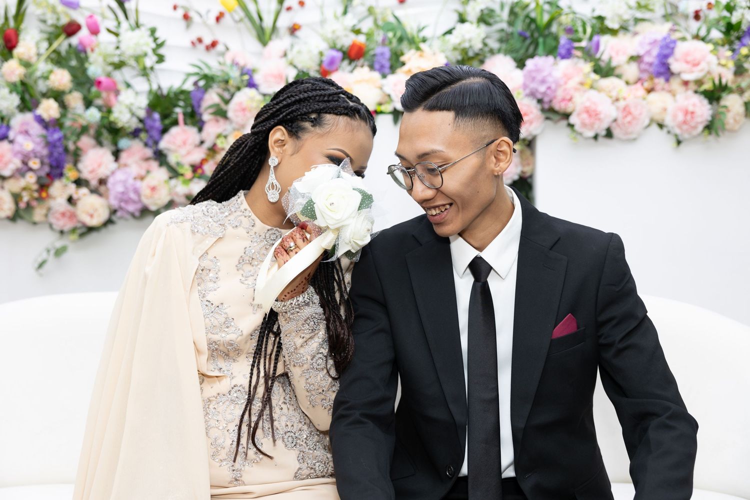 Couple in formal attire sharing intimate moment against floral wall backdrop.