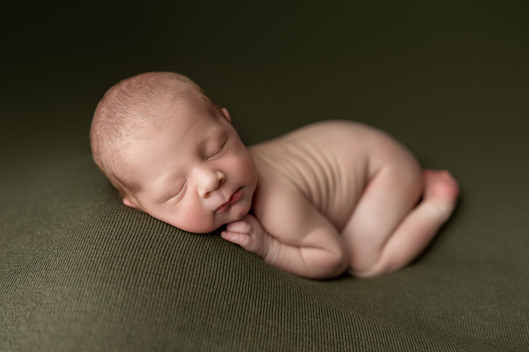 A sleeping newborn baby lies peacefully on a dark green textured backdrop in a professional photography setting.