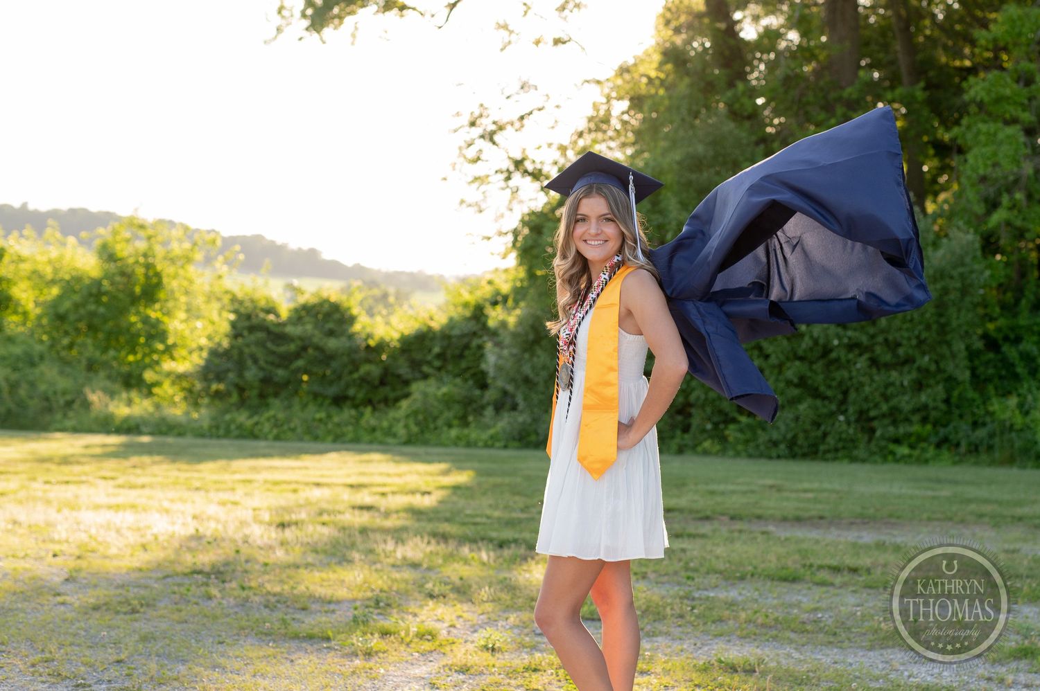Graduate in white dress and academic stole poses with gown flowing in evening light.