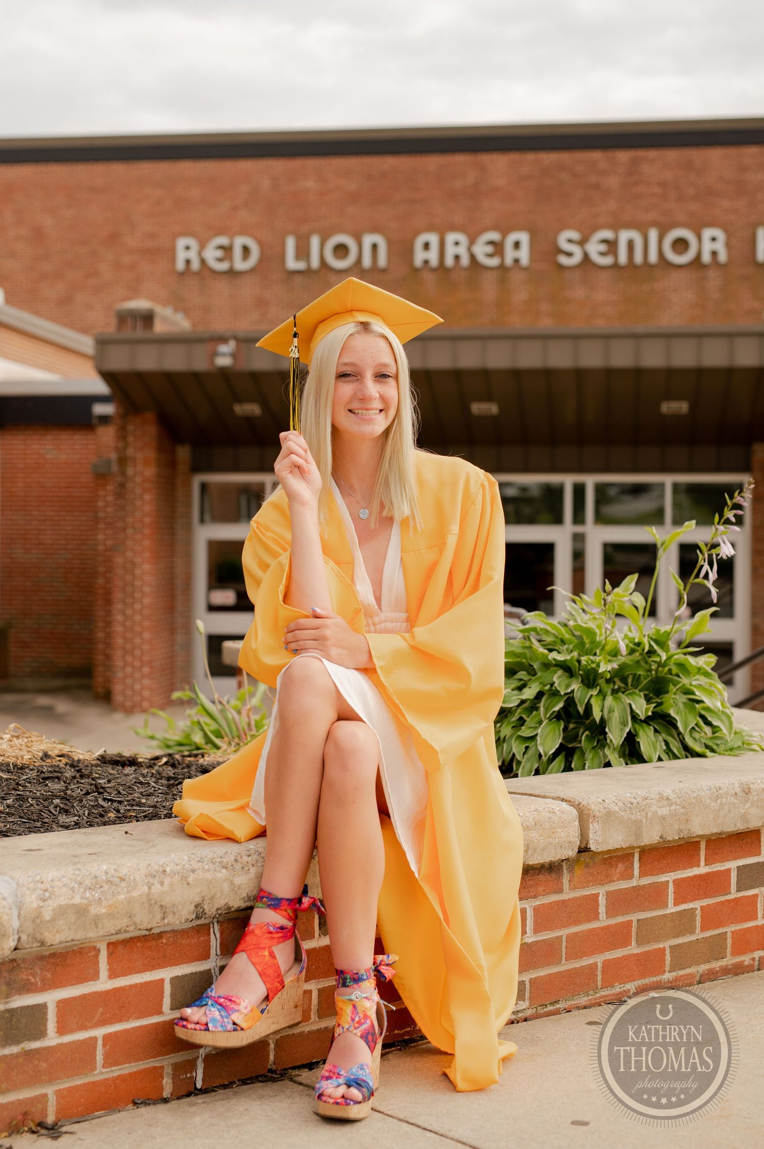 Graduate in yellow cap and gown sits on brick wall wearing colorful patterned shoes.