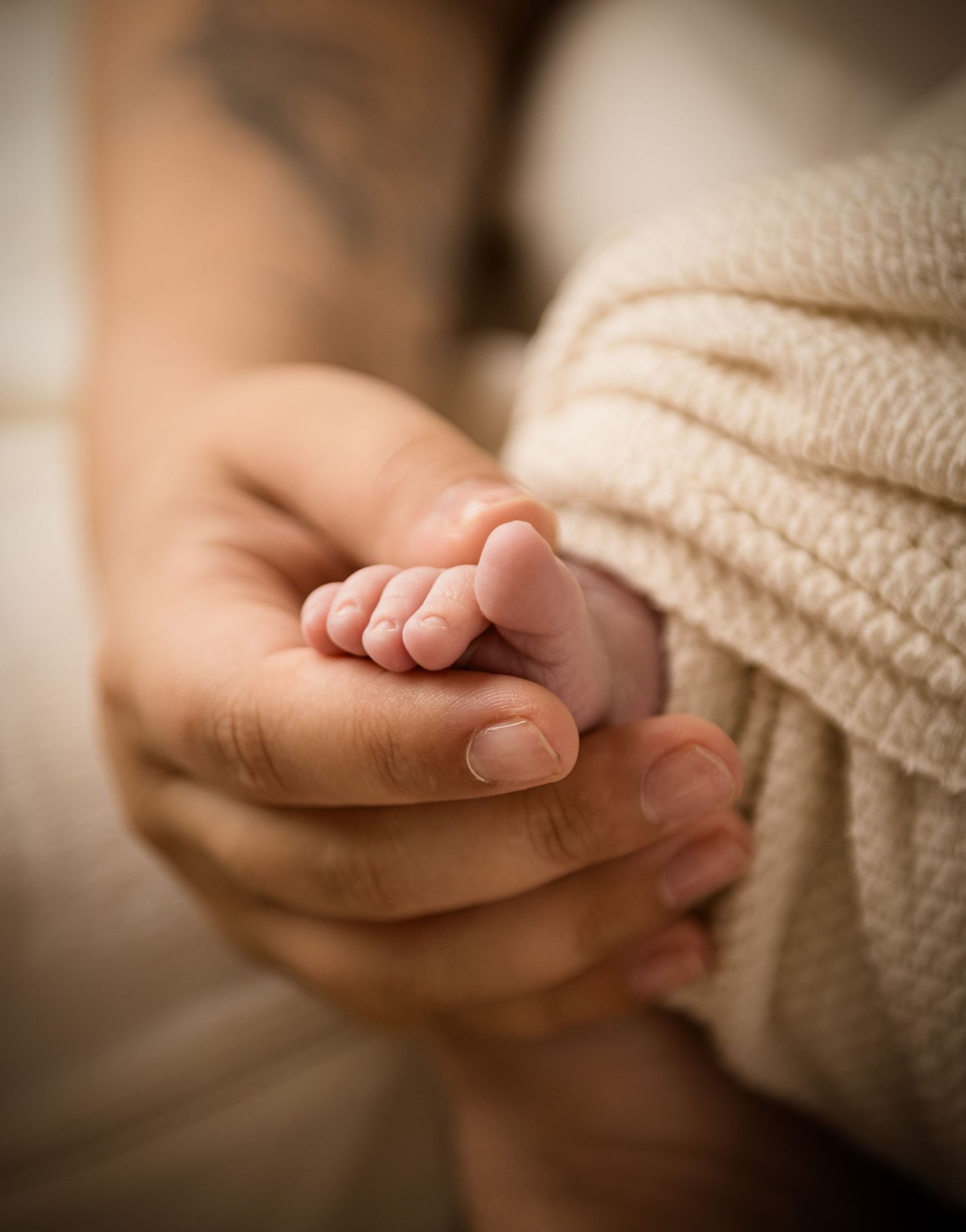 A tiny newborn baby foot rests gently in mum's protective hand against a soft cream knit blanket background.