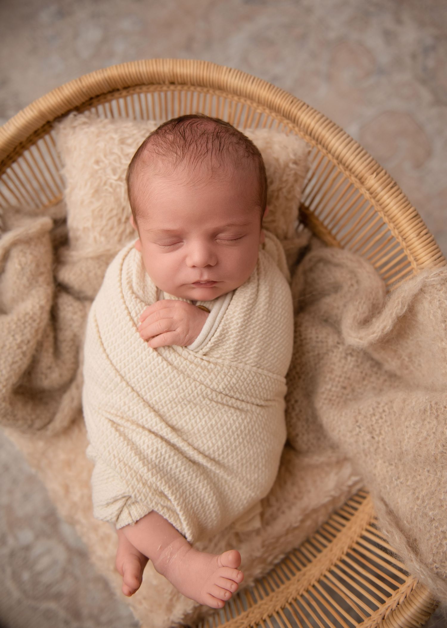 Sleeping newborn baby wrapped in cream knit blanket lying in woven basket with neutral toned background.