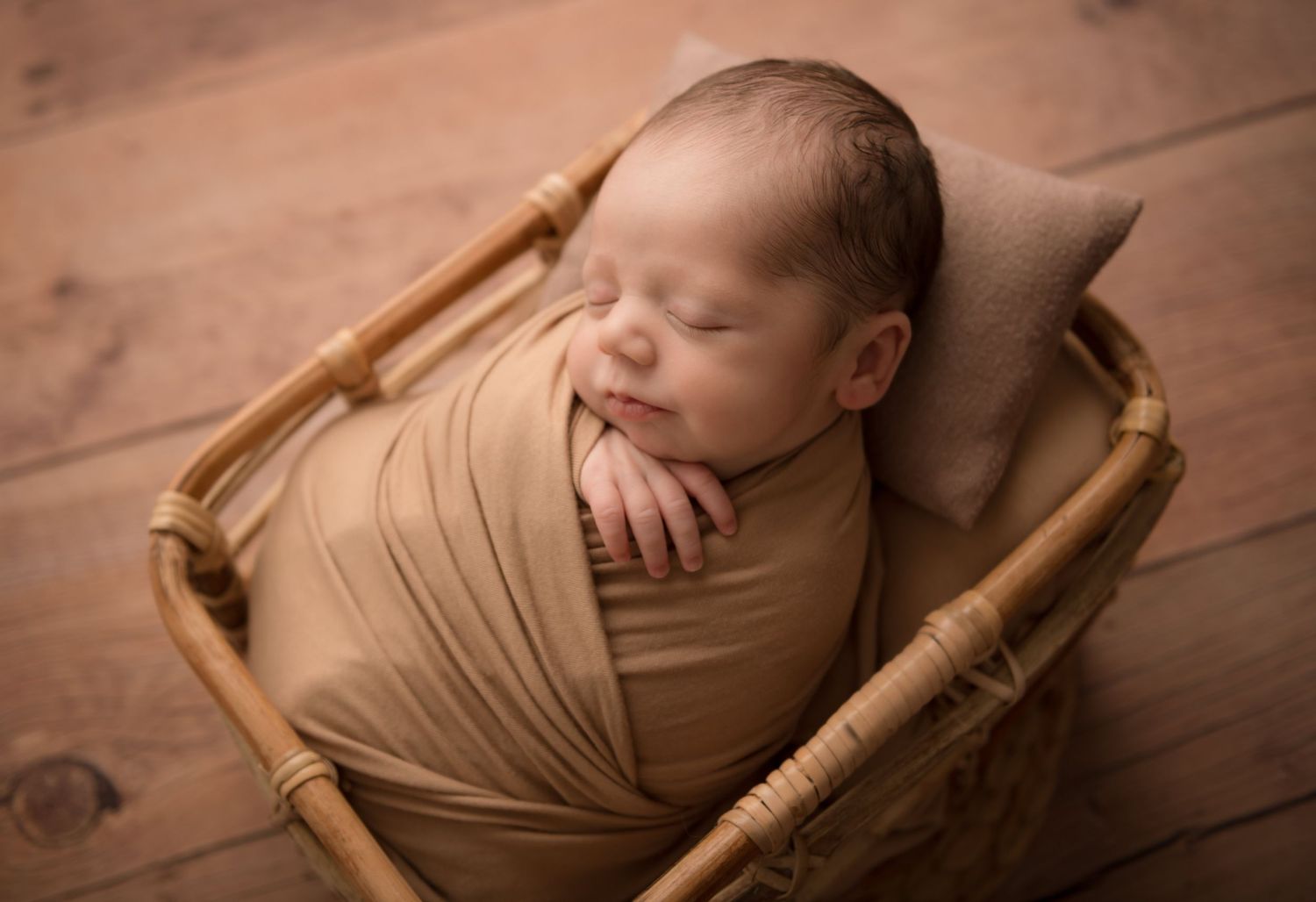 Peaceful sleeping newborn boy wrapped in tan fabric lying in a wooden basket.