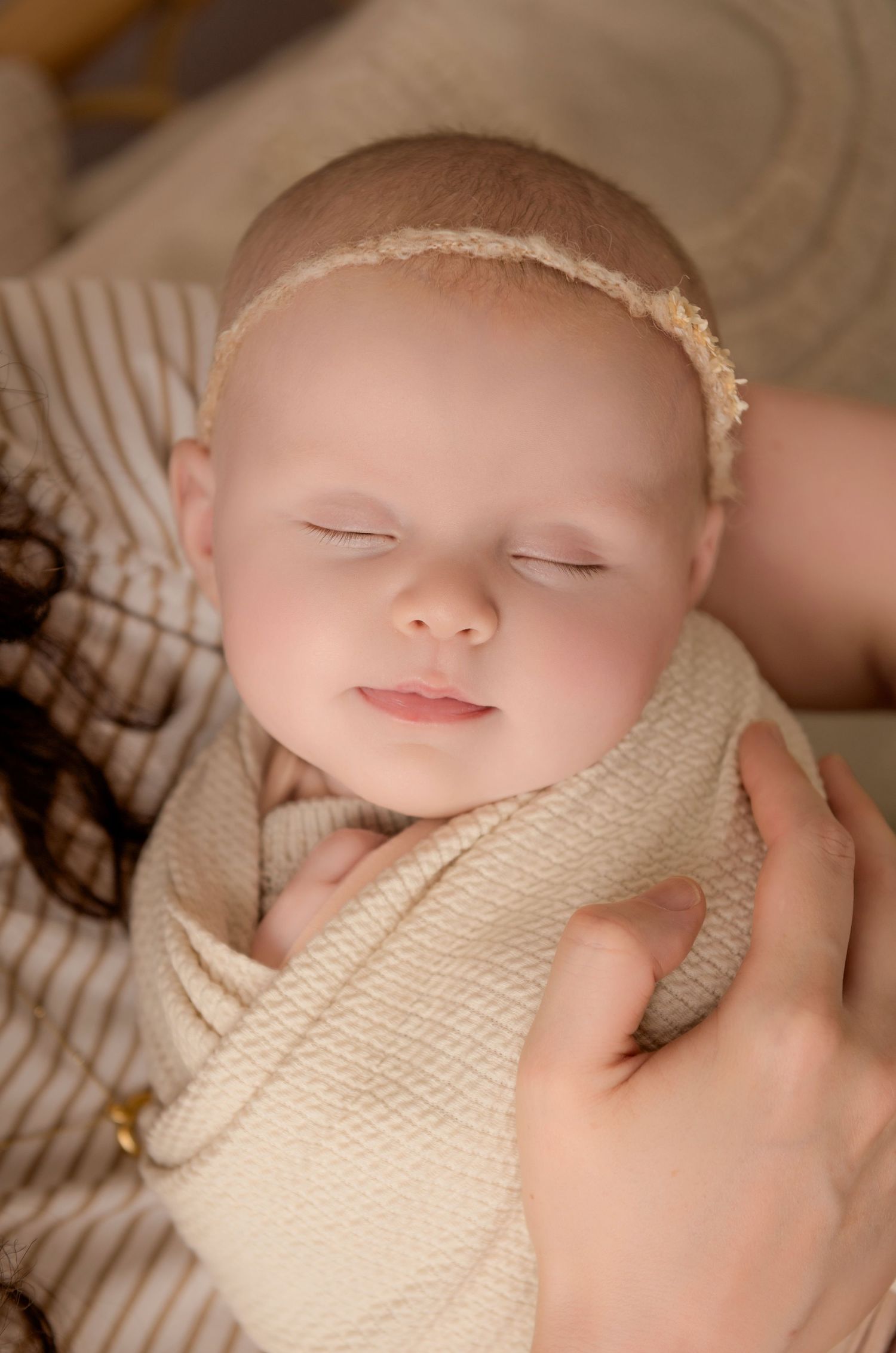 A sleeping newborn baby swaddled in cream-colored fabric with a delicate headband smiles in mum's arms.