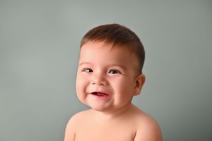 Smiling baby with dark hair and bright eyes against a soft sage green background, perfect for infant photography.