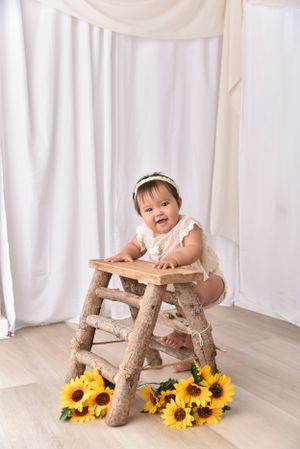 Baby in white dress sitting on rustic wooden stool surrounded by sunflowers.