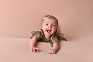 Joyful baby in olive green outfit laughing against peachy pink background.