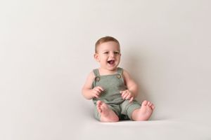 Happy baby in green romper sitting on white backdrop.