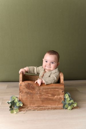 Baby wearing knit sweater sitting in rustic wooden box with greenery accents.