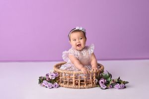 Professional baby photo shoot on purple backdrop with floral decorations and vintage wooden crate.