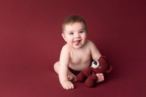 Baby photography session against deep red backdrop with plush toy companion.