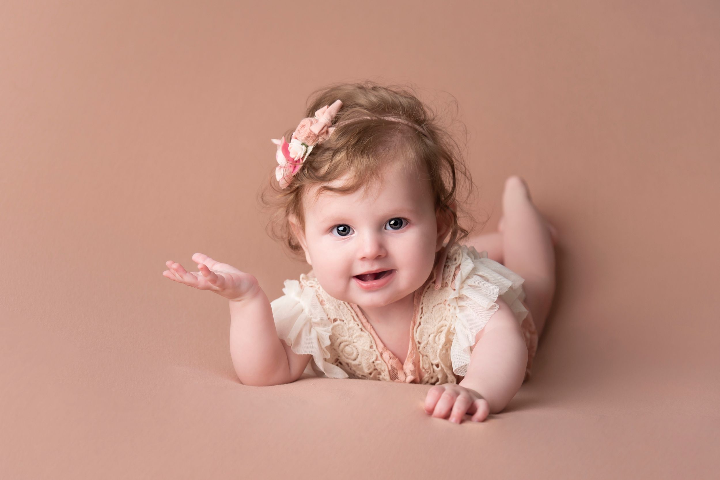 Baby in lace outfit smiling and reaching out against soft pink backdrop.