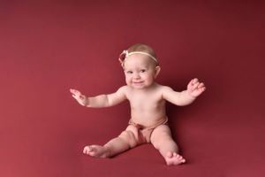 Happy baby sitting against burgundy backdrop with arms outstretched.