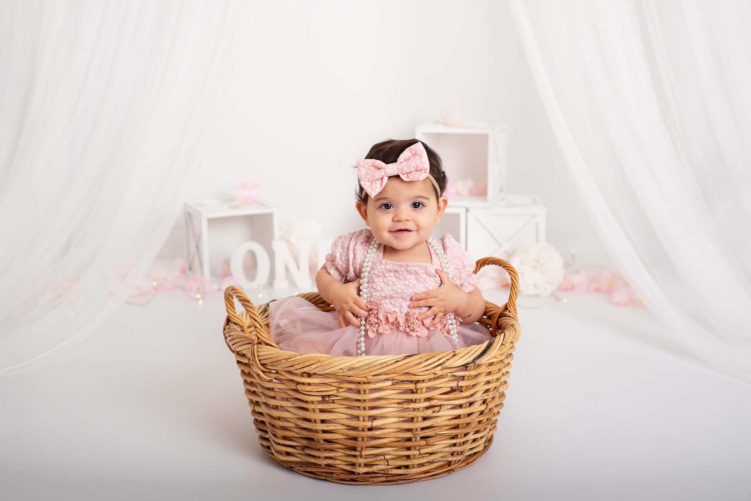 baby girl in pink sitting in basket for her one-year cake smash photo session, with soft white and pink decorations celebrating 1st birthday
