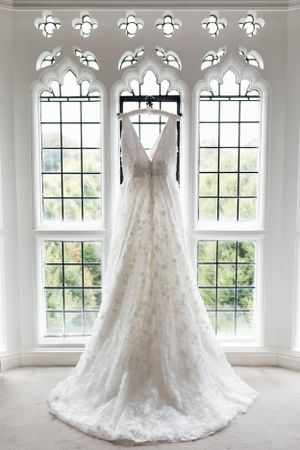 Elegant lace wedding dress hanging in front of gothic arched windows with natural light streaming through.