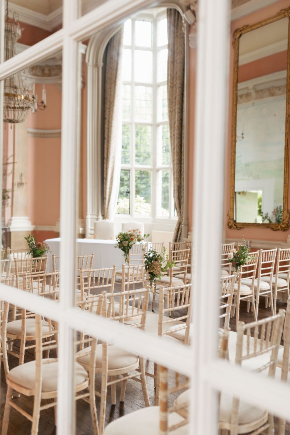 Elegant wedding venue with rows of white chiavari chairs and tall windows in a pink-walled room.