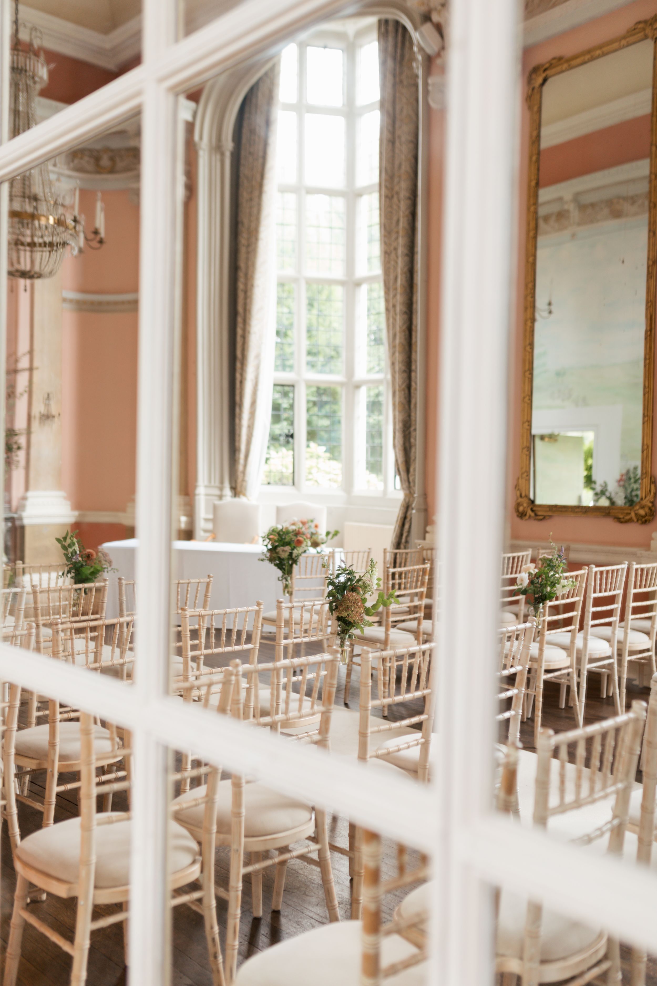 Elegant wedding venue with rows of white chiavari chairs and tall windows in a pink-walled room.