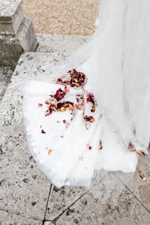 Red rose petals scattered on white wedding dress fabric against stone surface.