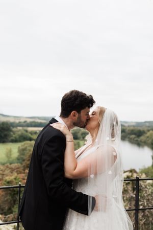 Romantic wedding couple sharing a kiss against scenic countryside river view.