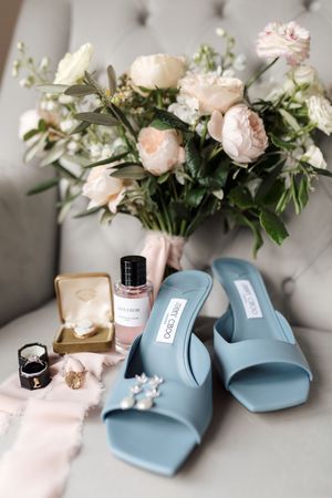 Elegant wedding details with light blue sandals, pink roses in a vase, and delicate jewelry on a white surface.