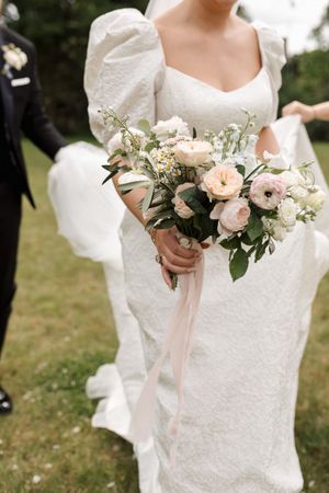 Delicate pink and white floral bouquet with trailing ribbons held against a white wedding dress.