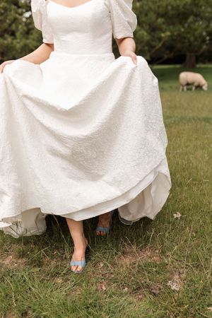 Close-up of white wedding dress hem and blue jimmy choo's on grassy ground, sheep in the background.