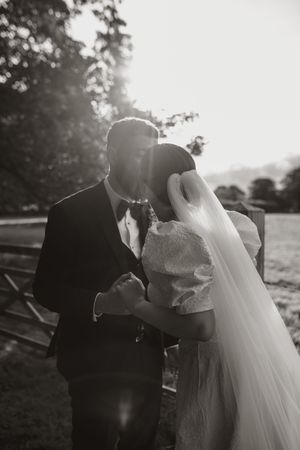 A romantic black and white wedding photo showing a couple embracing outdoors with sunlight filtering through trees.
