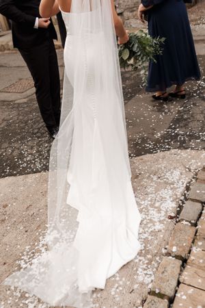 Long flowing white wedding veil and dress trailing on cobblestone ground with scattered flower petals.