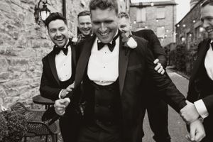 Black and white photo of groomsmen celebrating in formal attire against stone wall backdrop.