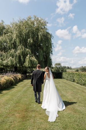 A couple walks hand in hand through a scenic countryside path lined with weeping willow trees on their wedding day.