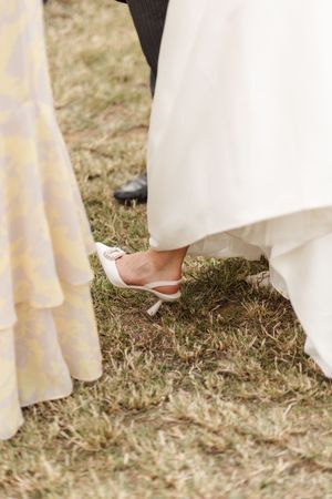 Close-up view of wedding dress hemlines and elegant flat shoes on grass during an outdoor ceremony.
