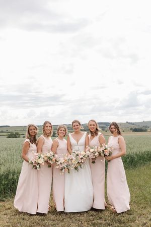 A group of bridesmaids in blush pink dresses pose together in a scenic field setting.