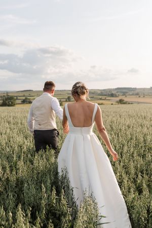 Couple walks through tall grass field in wedding attire during golden hour with rolling hills in background.