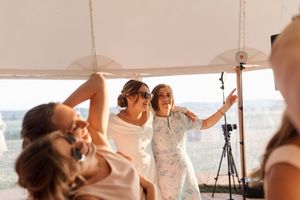 A bride in a white lace wedding dress gets her makeup professionally applied in a bright sunlit room.