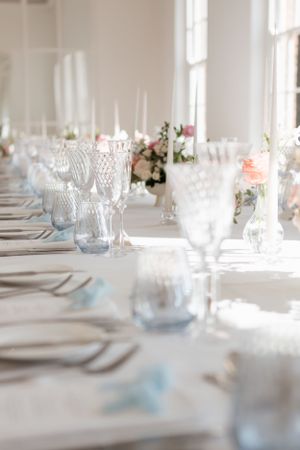 Elegant table setting with crystal glassware, white linens and soft pink floral centerpieces in natural sunlight.