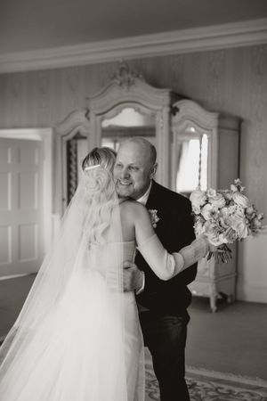 Emotional wedding moment captured in black and white with bride's veil and floral bouquet in elegant room.