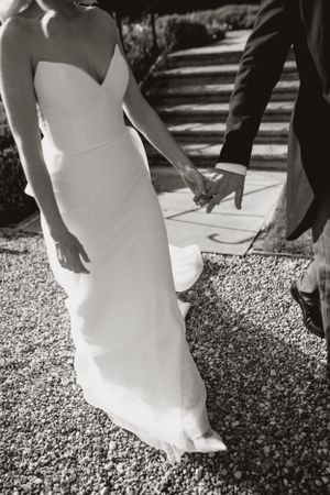 Black and white wedding photo showing couple holding hands, with bride in fitted strapless gown on gravel path.