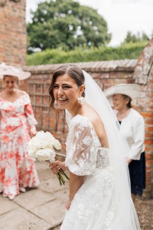 A joyful bride in a lace dress carries a white bouquet while walking through a brick-walled garden setting.