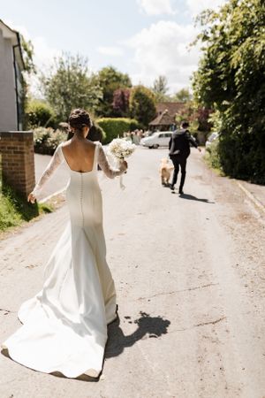 A bride in a flowing white dress walks down a country lane holding a bouquet.