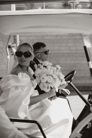 A classic black and white photo of newlyweds riding in a golf cart with a white rose bouquet.