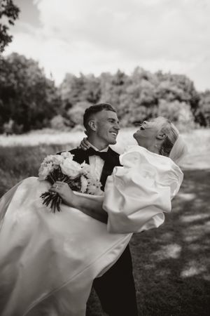 A joyful wedding moment captured in black and white as a groom carries his bride in a field.