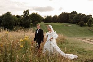 A newlywed couple walks through a scenic meadow with tall grass and trees in the background.