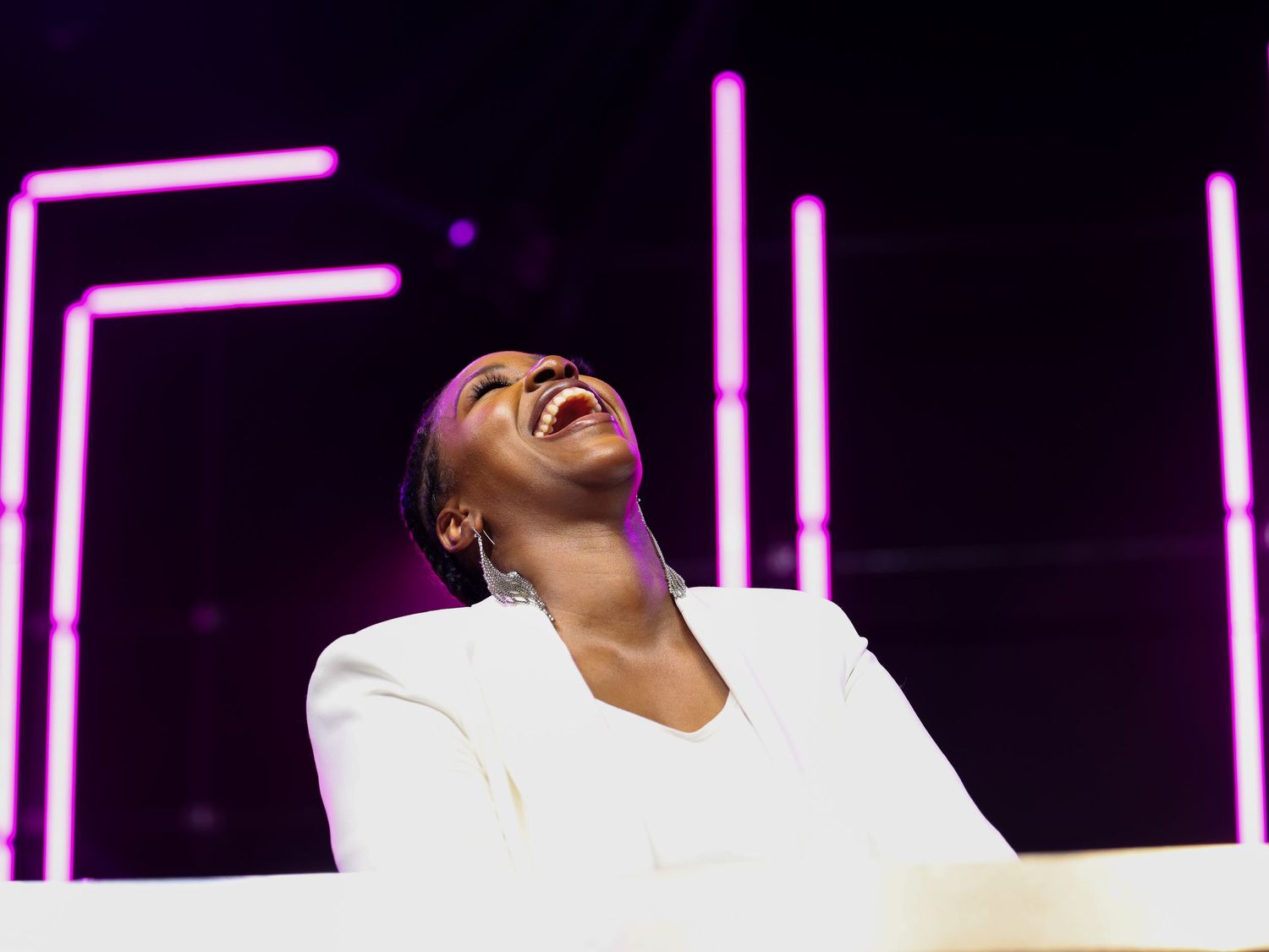 A performer in a white outfit laughs joyfully while performing on stage with pink neon lights in the background.