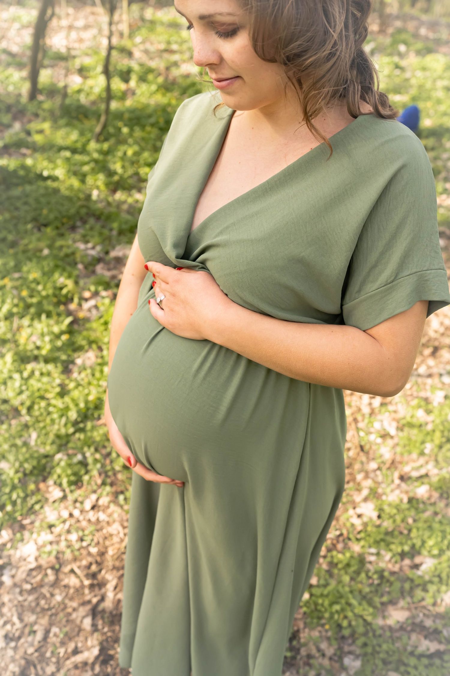 Close-up maternity portrait of woman in sage green dress amid spring forest backdrop.