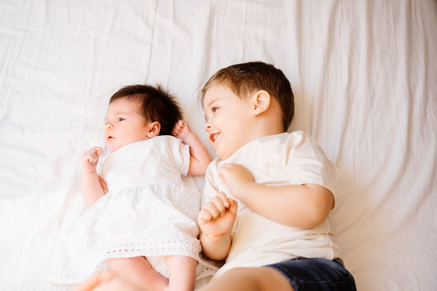 Two young children in white outfits lying together on white bedding.