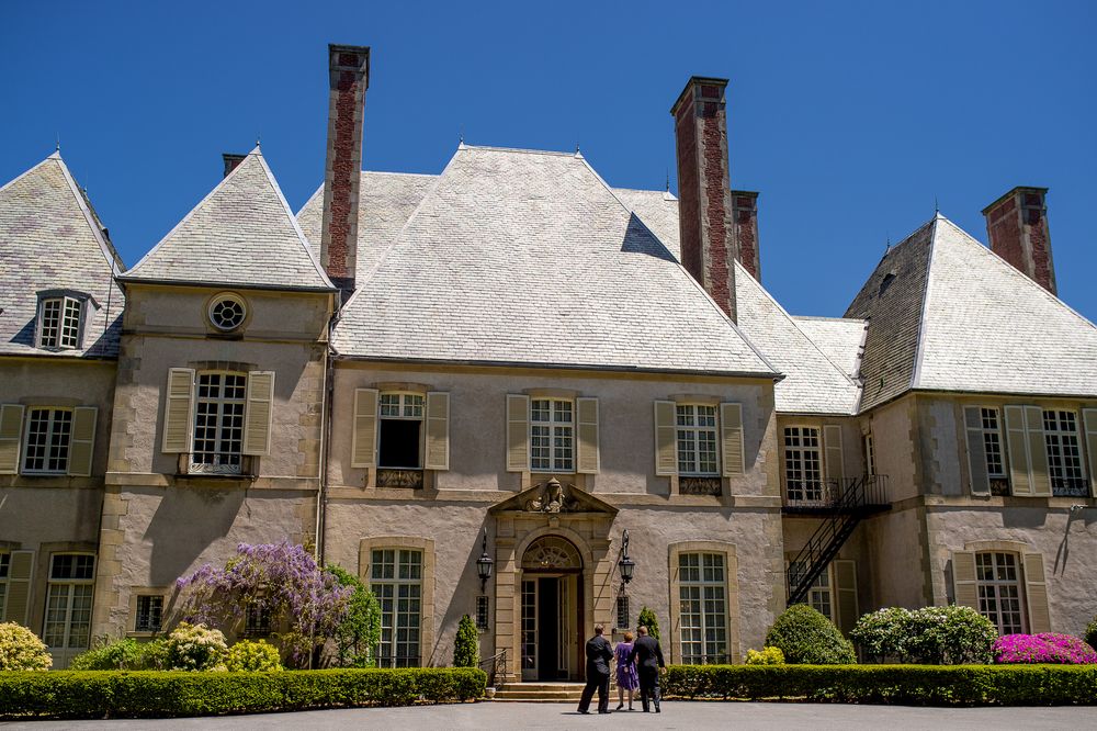 Large white estate mansion with multiple chimneys and manicured gardens on a sunny day.