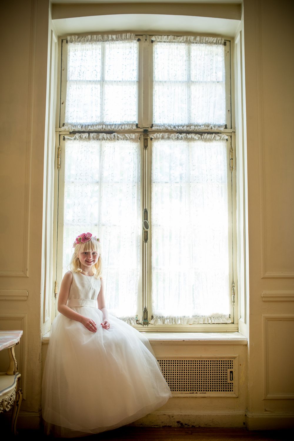 Bride in white wedding gown poses gracefully by sunlit window in vintage interior setting.