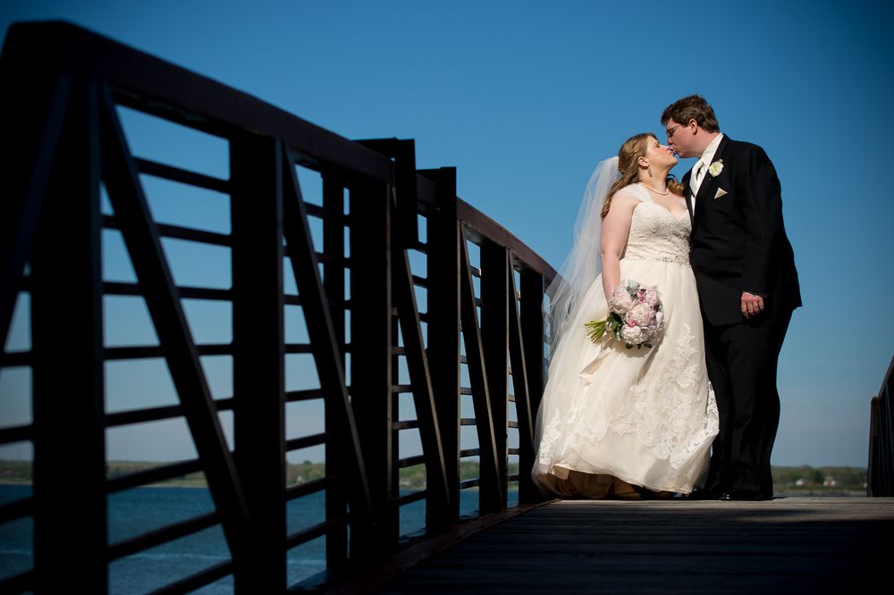 Silhouetted wedding couple embraces on wooden bridge pier against blue sky.