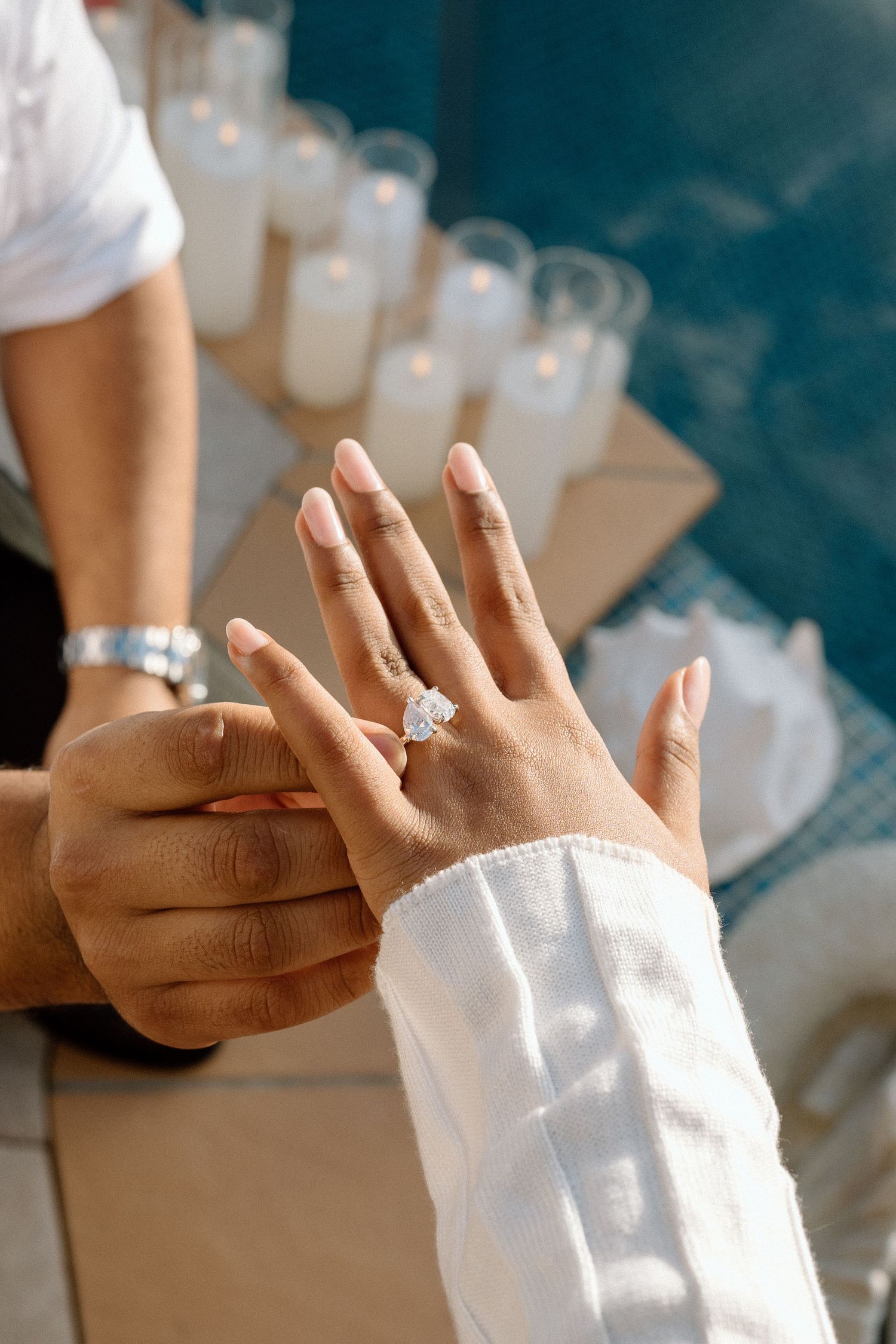 Close-up of hands showing off a sparkling engagement ring with water glasses in background.